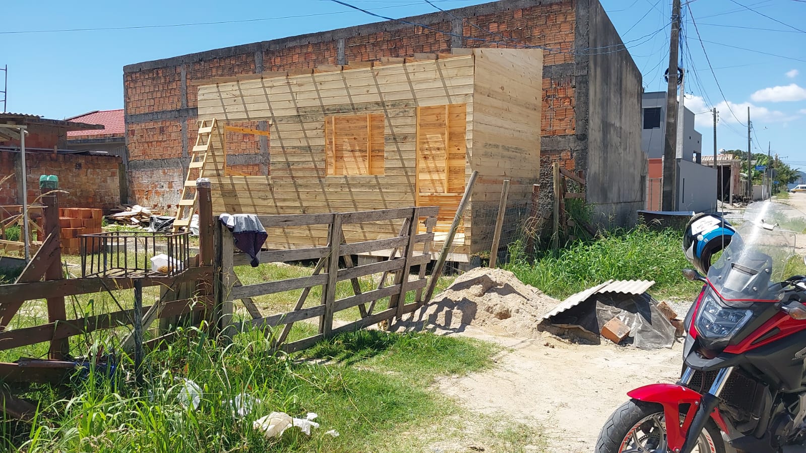 Construção de cabana de madeira em andamento, com uma moto e uma casa de alvenaria de fundo