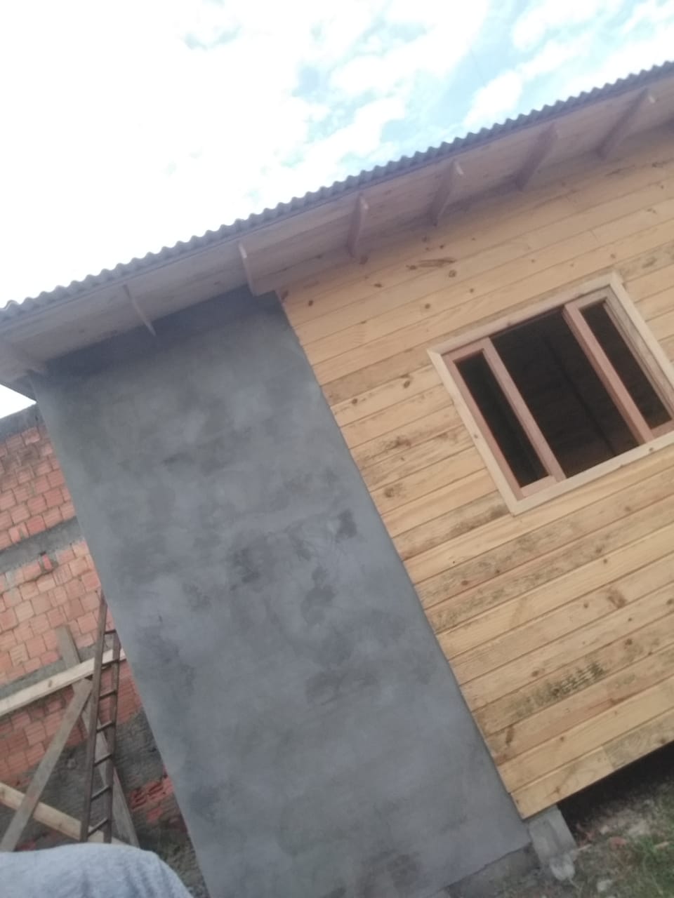 Construção em andamento, mostrando paredes de madeira e concreto, com uma janela e telhado em metal, e parte de uma parede de tijolo ao lado